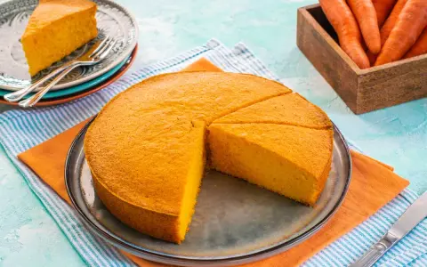 Una fetta di torta di carote sofficissima, appena tagliata da una torta intera su un piatto, con carote fresche in un cesto sullo sfondo.