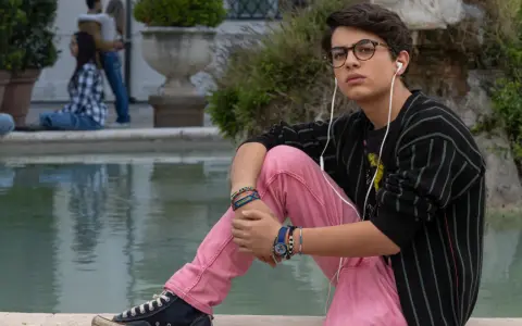 Il ragazzo dai pantaloni rosa, con occhiali e auricolari, ascolta musica seduto vicino a una fontana, mentre altre persone chiacchierano sullo sfondo.