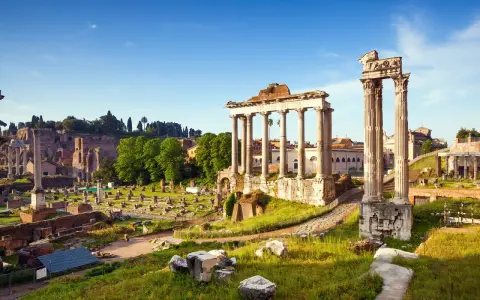 Antiche rovine del Foro Romano illuminate dal sole, con colonne imponenti e resti di templi sotto un cielo azzurro.