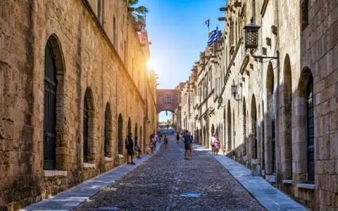 Sonnenbeschienene Gasse in Rhodos Stadt mit Kopfsteinpflaster und historischen Gebäuden. Touristen schlendern durch die mittelalterliche Stadt.