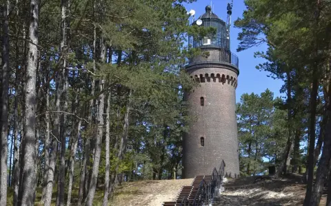 Ceglana latarnia Czołpino w lesie sosnowym, z drewnianymi schodami prowadzącymi do wejścia.