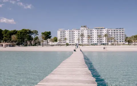 Drewniany pomost prowadzi do piaszczystej plaży Playa de Muro z białym hotelem i palmami w tle.