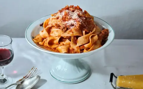 Ein Teller breiter Bandnudeln mit herzhaftem al ragu, bestreut mit Parmesan. Daneben ein Glas Rotwein und Besteck.