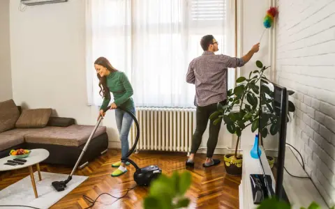 Pareja colaborando en como limpiar la casa: ella aspira el suelo y él quita el polvo de las paredes.