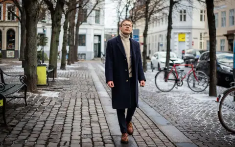 Elegancki mężczyzna w granatowym płaszczu damskim i golfie spaceruje po brukowanej ulicy. Stylowe granatowy płaszcz damski stylizacje.