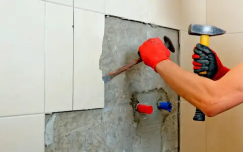 Man con guantes rojos usa un cincel y martillo para quitar azulejos y cemento de una pared.