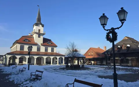 Zimowe miasteczko galicyjskie z ratuszem i karczmą, plac pokryty śniegiem, ławki i latarnie ozdobione świąteczną girlandą.