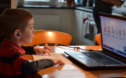 Chłopiec z niebieskim aparatem słuchowym ćwiczy wymowę sylab na lekcji logopeda online.