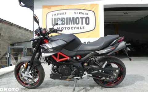 Czarny motocykl Aprilia Shiver 900 z czerwonym stelażem, zaparkowany przed salonem motocyklowym.