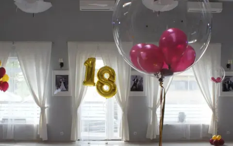 Złote balony z cyfrą 18 i przezroczyste balony z różowymi w środku ozdabiają salę. Balony na stołach tworzą radosną atmosferę.
