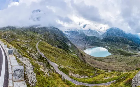 Strada tortuosa sul Passo San Lugano, con lago alpino azzurro e montagne nebbiose sullo sfondo.