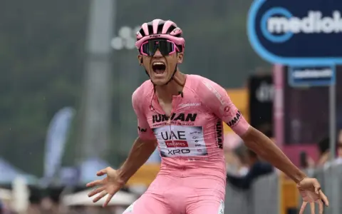 Isaac Del Toro, a cyclist in a pink jersey, screams in triumph, arms outstretched.
