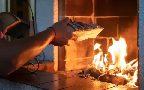 En hand lägger ved i en sprakande brasa. Det syns tecken på soteld i eldstaden.
