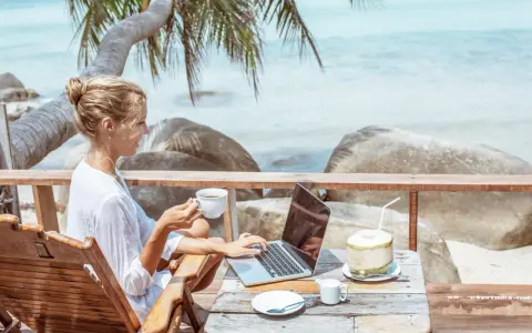 Kvinna med kaffe och laptop på en strandterrass, drömmen om ett digitalt nomad jobb.