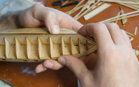 Hände bauen einen hölzernen Schiffsrumpf. Die Rippen des Kaders sind sichtbar, während die äußere Hülle geformt wird.