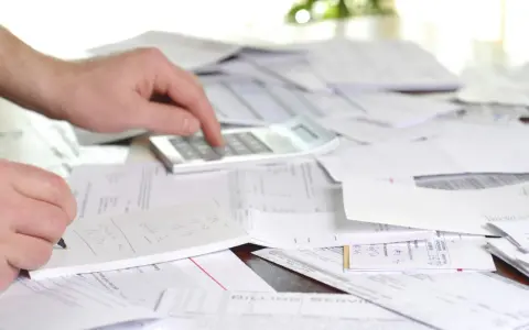 Mano calculando y anotando en libreta, rodeado de papeles y facturas con retención.