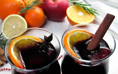 Dwa kieliszki z grzańcem, ozdobione plasterkiem pomarańczy i gwiazdką anyżu. Obok jabłka, pomarańcze, cytryna i laska cynamonu – idealne składniki, jak zrobić grzańca.