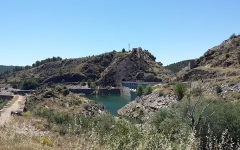 El embalse del Vado, con su presa de hormigón y aguas turquesas, se abre paso entre colinas rocosas y vegetación seca bajo un cielo azul.