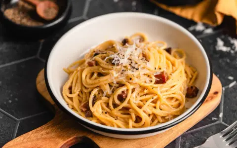 Eine Schüssel mit echter Carbonara, garniert mit geriebenem Parmesan und schwarzem Pfeffer. Daneben ein Holzlöffel mit Pfefferkörnern.