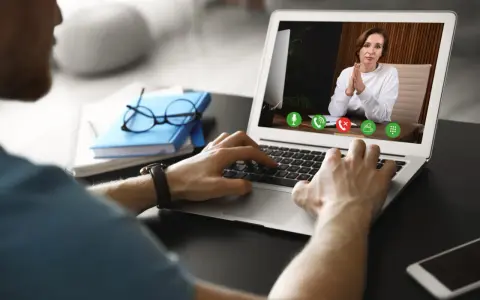 Mężczyzna prowadzi wirtualną superwizję przez laptopa. Na ekranie widać kobietę z dłońmi złożonymi w geście modlitwy.