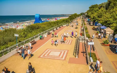 Tętniąca życiem ustka promenada w słoneczny dzień. Ludzie spacerują, odpoczywają na ławkach, a w tle widać plażę pełną turystów i morze.