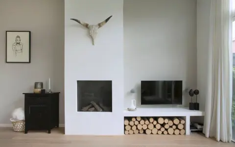 Un salon moderne avec une cheminée blanche épurée, un crâne de taureau, du bois de chauffage et une télévision. Un avant-après réussi pour relooker une cheminée.