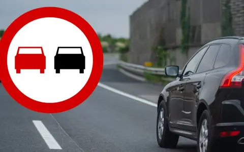 Znak zakazu wyprzedzania na podwójnej ciągłej. Czarny SUV jedzie po drodze, a na pierwszym planie znak ostrzegawczy.