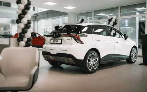 Białe auto MG 4 Electric w salonie, ozdobionym balonami. Obok czerwony samochód i wygodne fotele.