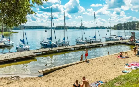 Słoneczny dzień nad jeziorami mazurskimi. Drewniane molo z żaglówkami, piaszczysta plaża z wypoczywającymi ludźmi i błękitne niebo z chmurami.