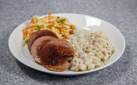 En tallrik med stuvade makaroner, stekt falukorv och en sallad på vitkål och morötter.