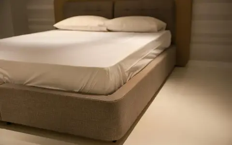Un lit moderne avec un sommier tapissier, une base recouverte de tissu beige, prêt à accueillir un matelas blanc et des oreillers.