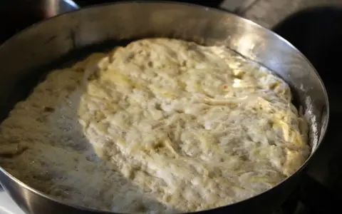 Hefeteig für Pizza geht auf. Die luftige Masse quillt über den Rand der Metallschüssel.