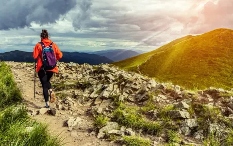 Kobieta w pomarańczowej kurtce wyrusza na szlak. To prawdziwy hiking po polsku, z pięknymi widokami i kamienistą ścieżką.