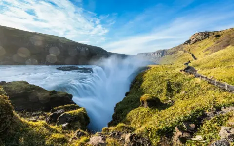 Potężny wodospad Gullfoss w Islandii, z mgiełką unoszącą się nad kaskadami. Ścieżka turystyczna wije się po zielonym zboczu.