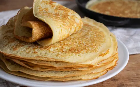 Pile de crêpes dorées, une roulée sur le dessus. Une poêle avec une crêpe en cuisson est visible en arrière-plan. Parfait pour 1 litre de pâte à crêpe.