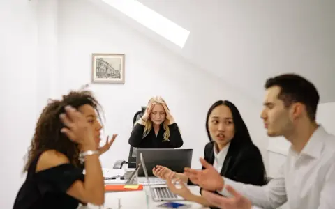 Un gruppo di colleghi di lavoro discute animatamente. Una donna bionda sembra stressata, tenendosi la testa tra le mani.