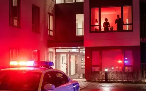 Radiowóz policyjny z włączonymi sygnałami świetlnymi przed budynkiem mieszkalnym. W oknie widać sylwetki ludzi, być może naruszających ciszę nocną w Polsce.