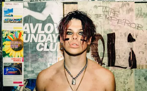 Yungblud, "The Emperor", z charakterystycznym makijażem i łańcuchami, na tle plakatów.