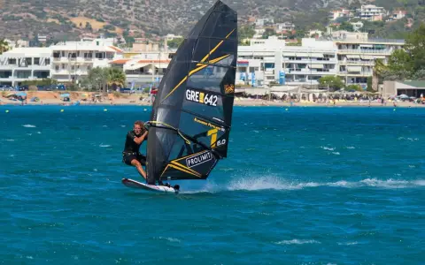 Ein Mann beim Windsurfen in Griechenland, mit Blick auf eine belebte Küste und Berge im Hintergrund.
