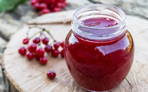 Un pot de gelee de groseille, aux reflets rubis, est posé sur une planche de bois. Des grappes de groseilles rouges sont disposées à côté.