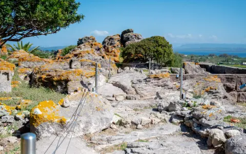 Ancient Italian ruins with weathered stones covered in orange lichen, a pathway winding through the site, and a distant blue sea under a clear sky.