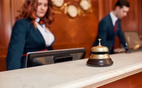 Recepcjonistka i recepcjonista za ladą hotelową. Dzwonek do obsługi gości, który pomoże dowiedzieć się, ile kosztuje doba hotelowa.