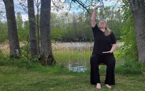 Kvinna i svart klädsel utför yoga på en stol vid en sjö. Naturen är grönskande med träd och vass.