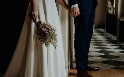 Para młoda trzyma się za ręce, ona z bukietem suszonych kwiatów. Wesele motywy w eleganckim wnętrzu.