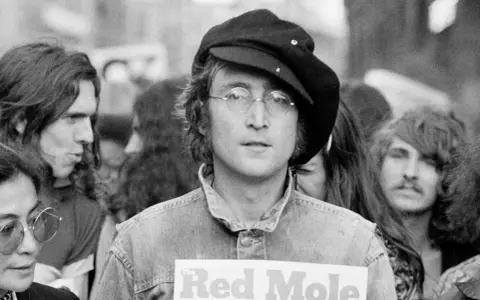John Lennon w czapce i okularach, trzymający plakat "Red Mole". W tle inni ludzie, być może fani jego utworów.