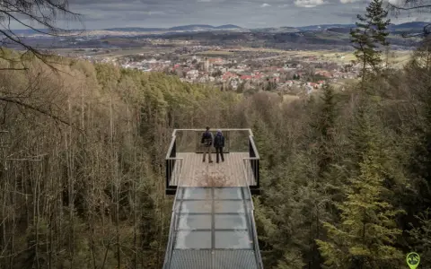 Dwie osoby stoją na szklanym tarasie widokowym, podziwiając panoramę Starego Sącza i okolic.