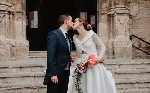 Novios besándose frente a una iglesia, ella lleva un ramo de flores. Un momento especial tras las lecturas de boda religiosa.