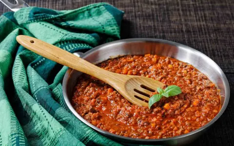 Un saporito ragù di cinghiale in padella, pronto per condire la pasta. Un cucchiaio di legno e una fogliolina di basilico completano la scena.