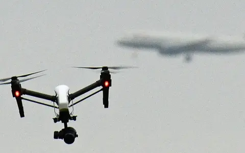 A white drone with red lights hovers in the sky, possibly on a security mission, as a blurred airplane flies in the distance.