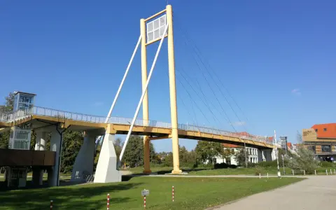 Żółty most pieszy, molo w Giżycku, z charakterystycznymi pylonami i linami, prowadzi do budynków nad jeziorem.